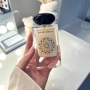 Hand holding a perfume bottle labeled 'Musk Collection Tahara Vanilla' on a blurred background
